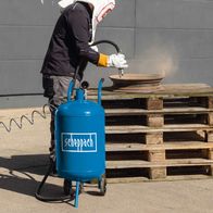 Mobiles Sandstrahlgerät SBT-80 von Scheppach. Ein Arbeiter verwendet das Gerät zum Sandstrahlen von Holzpaletten. Das Gerät verfügt über einen blauen Tank mit dem Markennamen Scheppach und einen Schlauch mit einer Handstrahlpistole.