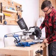 Ein Mann bedient eine Kappzugsäge HM254 von Scheppach in einer Werkstatt. Die Säge steht auf einer Holzbank und ist mit einem Holzbrett im Einsatz. Im Hintergrund sind Werkzeuge an der Wand befestigt.