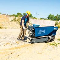 Kettendumper DP5000 Scheppach im Einsatz auf einer Baustelle. Ein Arbeiter in Schutzausrüstung bedient das Gerät auf einem unebenen Gelände. Scheppach-Logo sichtbar auf dem Behälter.