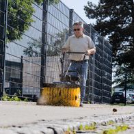 Ein Mann bedient die Kehrmaschine SC2400p von Scheppach auf einem Gehweg. Die Maschine ist in Betrieb und wirft Schmutz auf. Im Hintergrund ist ein Zaun und ein Auto zu sehen.