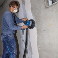 Ein Arbeiter nutzt den Wand- und Deckenschleifer DS200 von Scheppach an einer Wand. Der Schleifer hat einen Durchmesser von 215 mm und eine Leistung von 1200 W. Der Arbeiter trägt Schutzausrüstung.