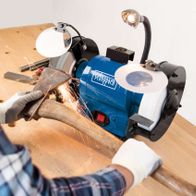 Ein Mann nutzt den Doppelschleifer BG200AL von Scheppach, um Metall zu bearbeiten. Der Schleifer ist auf einem Holztisch montiert und wird mit beiden Händen gehalten. Der Mann trägt eine Schutzbrille und Handschuhe.