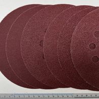 Ein Stapel aus zehn runden Schleifpapieren mit verschiedenen Körnungen, gemessen an einem Lineal. Die Scheiben haben Löcher für eine Schleifmaschine.