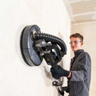 Ein Mann verwendet den Scheppach Wand- und Deckenschleifer DWS90X Woodster an einer Wand. Der Schleifer hat eine Teleskopfunktion und eine LED-Leuchte.