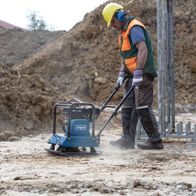 Ein Arbeiter bedient eine Scheppach Rüttelplatte HP1800S auf einer Baustelle. Die Maschine ist blau und hat einen orangefarbenen Sicherheitsvest.