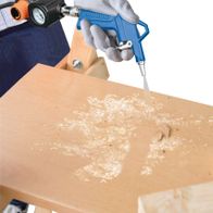 Ein Scheppach Kompressor wird verwendet, um Holz zu bearbeiten. Der Kompressor wird von einer Hand gehalten und sprüht Luft auf ein Holzbrett, das auf einer Werkbank liegt. Holzspäne sind sichtbar.