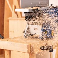 Ein Scheppach SprayVac20-Sauger saugt Holzspäne von einer Werkbank. Im Hintergrund ist eine Kreissäge zu sehen.