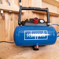 Scheppach air compressor HC06 with accessories on a wooden table. The compressor has a blue tank and is connected with a blue hose.