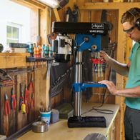 A man operates a Scheppach bench pillar drill DP19Vario in a workshop. The man is wearing safety goggles and ear protection. The workbench is equipped with tools and accessories.