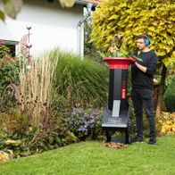 Ein Mann bedient den Scheppach BIOSTAR 3000 Häcksler im Garten. Der Häcksler steht auf einem Rasenstück umgeben von Pflanzen und Laub.