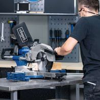 Ein Mann bedient eine Scheppach-Kappzugsäge HM110MP in einer Werkstatt. Die Säge steht auf einer Metallplatte und hat eine schwarze Schutzhaube. Im Hintergrund sind Werkzeuge an der Wand aufgehängt.