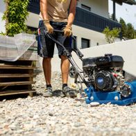 Rüttelplatte HP1900S von Scheppach im Einsatz, Person bedient die Maschine auf einem Kiesboden. Die Maschine hat einen schwarzen Motor mit blauem Gehäuse.