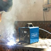 Ein Arbeiter schweißt mit dem 3in1 Kombi-Schweißgerät WSE4000-CUT von Scheppach. Der Schweißer trägt Schutzausrüstung und arbeitet an einem Metallgitter. Das Gerät steht auf dem Tisch und erzeugt Rauch.