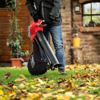 Ein Mann verwendet den Laubsauger und -bläser BBV500-40Li von Scheppach, um Blätter auf einer Wiese aufzusaugen. Der Laubsauger und -bläser ist schwarz und rot. Im Hintergrund befindet sich ein Haus mit einem Fenster und Pflanzen.
