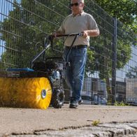 Ein Mann bedient eine Kehrmaschine SC2400p von Scheppach auf einem Gehweg. Die Maschine hat eine gelbe Bürste und wirft Laub zur Seite.