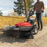 Die Kehrmaschine SC2200PE von Scheppach mit Sammelbehälter 70l und Schneeschild. Die Maschine ist im Einsatz zu sehen, wie sie Laub und kleine Äste auf einer Straße aufnimmt.