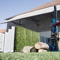 Ein Mann bedient einen Meterholzspalter HL3000GM von Scheppach. Er trägt Schutzausrüstung und schiebt einen Holzstamm in die Maschine. Im Hintergrund befindet sich ein Gebäude mit einer Hecke. Der Holzspalter ist blau und weiß.