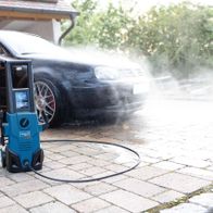 Scheppach Hochdruckreiniger HCE1600 im Einsatz, ein Mann reinigt ein Auto auf einem Hof mit Kopfsteinpflaster.