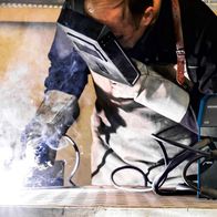 Ein Arbeiter schweißt mit dem Multischweißgerät WSE4000 von Scheppach. Der Schweißer trägt Schutzausrüstung und arbeitet an einem Metallstück. Der Schweißprozess erzeugt Rauch.