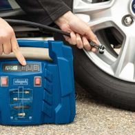 Ein Scheppach Kompressor mit LCD-Display und Druckanzeige. Eine Hand hält den Kompressor, während die andere ein Werkzeug hält. Der Kompressor ist mit einem Auto verbunden.