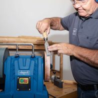 A man is sharpening a knife using the Scheppach Wet Bench Grinder Tiger 7000S. He holds the knife against the rotating grinding wheel, carefully guiding it along the blade.