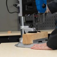 Tischbohrmaschine DP60 Scheppach im Einsatz. Ein Holzblock wird auf der Arbeitsplatte fixiert und mit einem Bohrer bearbeitet.