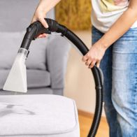 Ein Scheppach SprayVac20-Sauger entfernt einen Fleck von einem hellen Sofa. Die Person hält den Sauger mit beiden Händen und saugt den Fleck auf.