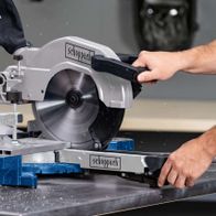 Kappzugsäge HM110MP von Scheppach im Einsatz. Die Säge schneidet ein Material, während ein Benutzer sie bedient. Die Säge ist auf einem blauen und silbernen Gestell montiert.