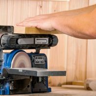 Band- und Tellerschleifer BTS900 von Scheppach. Ein Holzblock wird auf der Scheibe geschliffen. Die Maschine ist von der Seite zu sehen.