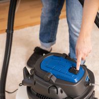 Ein Scheppach-Sprüh- und Waschsauger mit blauer Abdeckung und schwarzem Griff. Eine Hand drückt den Einschaltknopf.