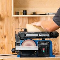 Band- und Tellerschleifer BTS900 von Scheppach im Einsatz. Ein Nutzer schleift ein Holzbrett an der Scheibe. Die Maschine ist auf einem Holzregal montiert.