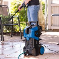 Der Scheppach Hochdruckreiniger HCE3000 mit 2400W, 180bar, 8m Anti-Twist Schlauch und 480 L/h Fördermenge. Gezeigt wird der Reiniger von der Seite mit einem Gartensitzmöbel im Hintergrund.