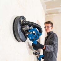 Wand-Deckenschleifer DS920X von Scheppach im Einsatz. Ein Arbeiter nutzt das Gerät an einer Wand. Der Schleifer hat einen großen runden Kopf und einen flexiblen Schlauch.