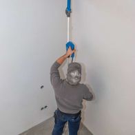 Ein Arbeiter verwendet den Wand- und Deckenschleifer DS930 von Scheppach an einer Wand. Der Schleifkopf ist in Aktion und entfernt Material von der Oberfläche. Der Arbeiter trägt Schutzausrüstung, einschließlich einer Maske und Handschuhen.