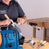 Ein Mann bedient eine Scheppach Nass-Schleifmaschine. Er hält einen Holzgriff in der Hand und justiert die Maschine. Im Hintergrund ist eine Werkbank mit verschiedenen Werkzeugen zu sehen.