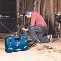 Ein Mann kniet auf dem Boden und repariert ein Fahrrad. Neben ihm steht ein Scheppach Kompressor. Im Hintergrund sind Werkzeuge und Materialien zu sehen.