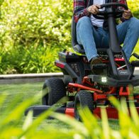 Akku-Aufsitzrasenmäher MR-760E von Scheppach im Einsatz. Person sitzt auf dem Mähgerät und steuert es auf einem Rasen. Das Gerät hat LED-Licht und einen Tempomat.