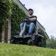 Mann auf Scheppach-Aufsitzrasenmäher mit LED-Licht und Abdeckhaube, zeigt Produkt mit 61 cm Schnittbreite und 150-Liter-Korb
