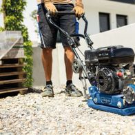 Ein Mann bedient eine Scheppach Rüttelplatte HP1300S auf einem Kiesboden. Die Maschine hat einen schwarzen Motor und einen blauen Rahmen. Der Name 'Scheppach' ist auf dem Gehäuse sichtbar.