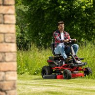 Ein Mann sitzt auf einem Akku-Aufsitzrasenmäher MR-760E von Scheppach und mäht einen Rasen. Der Mäher hat eine Schnittbreite von 76 cm und ist mit LED-Licht und Tempomat ausgestattet.