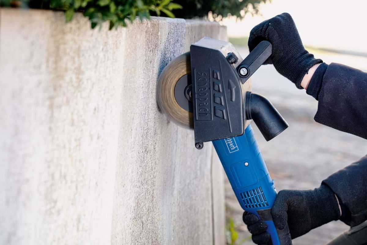 Scheppach Messingdrahtbürste MRS1300 im Einsatz an einer Wand. Die Bürste ist an einem Winkelschleifer befestigt und wird von einer Person mit Handschuhen gehalten.