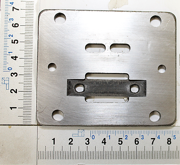 Ventilplatte mit Messskala im Hintergrund. Die Platte hat mehrere Löcher und Aussparungen. Die Platte wird von der Seite gezeigt.