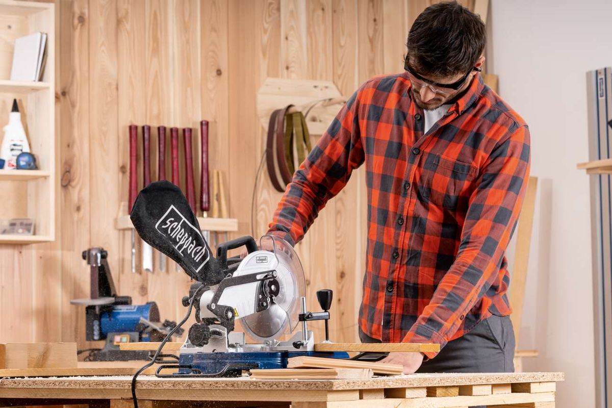 Ein Mann bedient eine Kapp- und Gehrungssäge HM80L von Scheppach in einer Werkstatt. Die Säge ist auf einem Holztisch montiert und schneidet durch ein Holzbrett. Im Hintergrund sind verschiedene Werkzeuge und Materialien zu sehen.