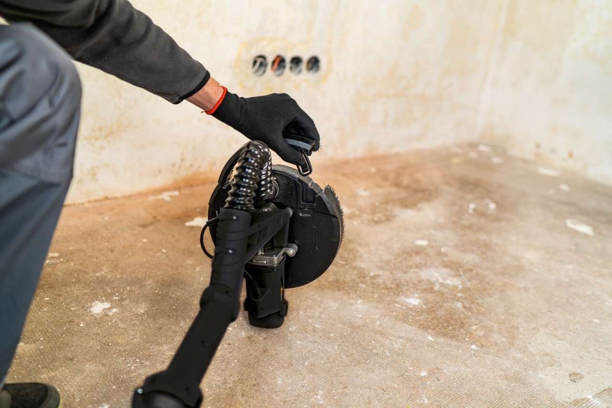 Ein Mann hält den Scheppach Wand- und Deckenschleifer DWS90X Woodster in der Hand. Der Schleifer ist auf den Boden gerichtet.