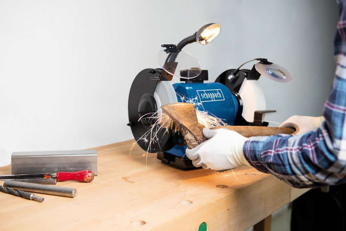 Scheppach Doppelschleifer BG200AL im Einsatz. Ein Benutzer schleift ein Holzstück mit dem Gerät. Werkzeuge und Schleifblöcke liegen auf dem Tisch.