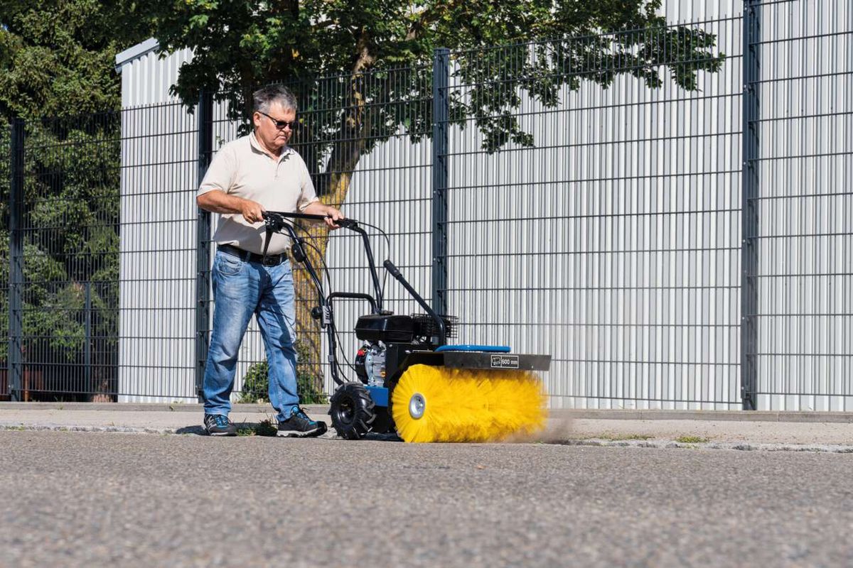 Ein Mann bedient eine Kehrmaschine SC2400p von Scheppach auf einem asphaltierten Weg. Die Maschine hat einen gelben Besen und einen Auffangbehälter.
