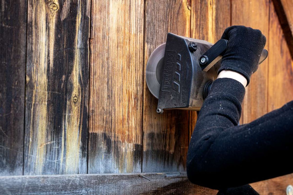 Scheppach Schleiflamellenbürste im Einsatz an einer Holzoberfläche. Die Bürste wird von einer Hand mit Handschuh gehalten und bearbeitet eine Holzwand.