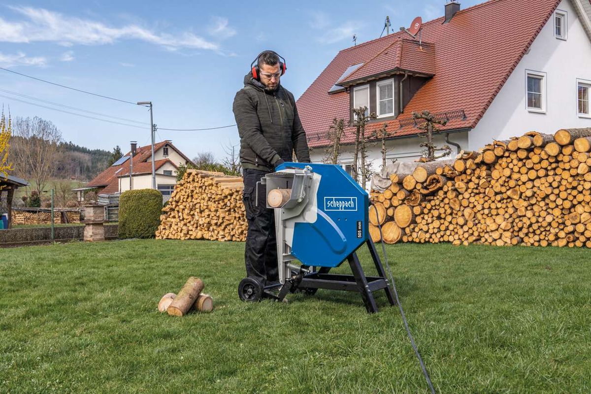 Ein Mann bedient die Wippsäge HS510 von Scheppach im Garten. Im Hintergrund sind Holzstapel und ein Haus zu sehen.