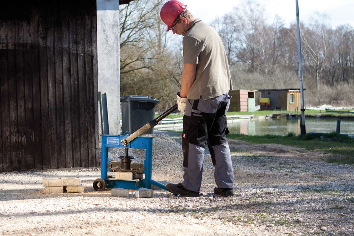 Ein Arbeiter bedient einen Steinknacker HSC130 von Scheppach. Der Mann trägt eine rote Schutzhelm und Arbeitskleidung. Im Hintergrund ist ein Gewässer und eine kleine Hütte zu sehen.