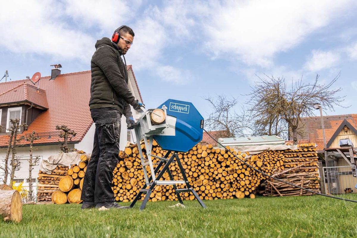 Ein Mann bedient eine Wippsäge HS410 von Scheppach im Garten. Im Hintergrund sind Holzstapel zu sehen.